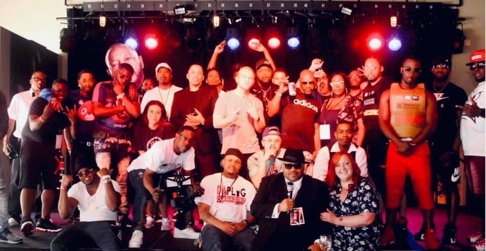 Group of people on stage with colorful lights, some holding microphones, posing for a photo.