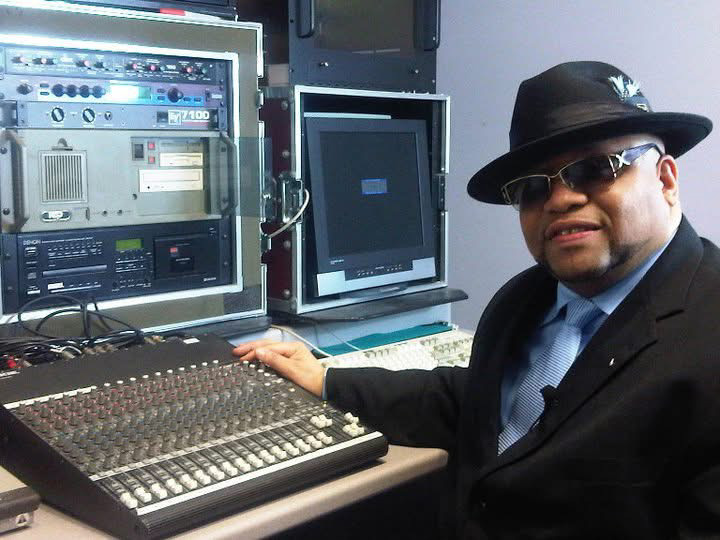 Man in black hat and sunglasses sitting at a mixing console with computer screens behind him.
