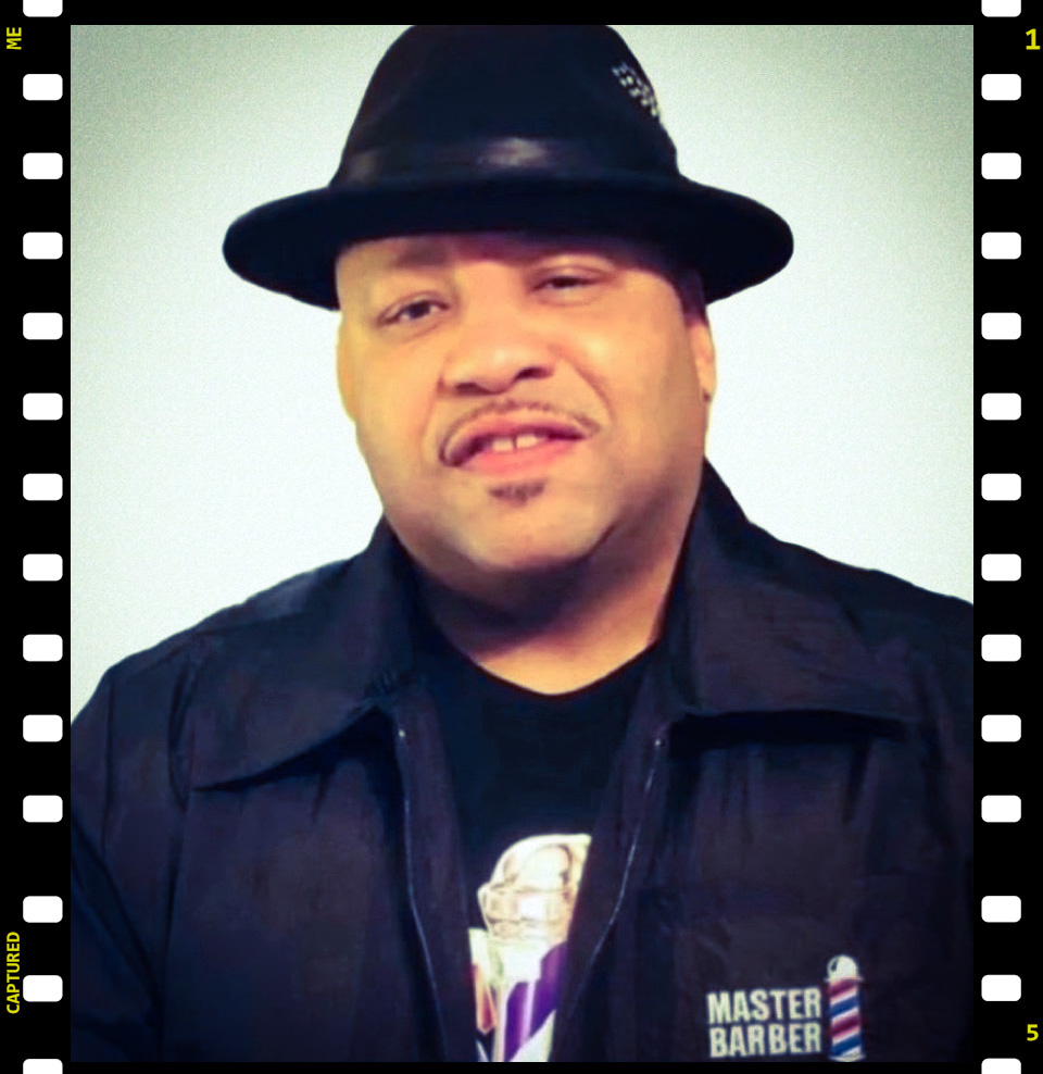 Person wearing a black hat and jacket, making a facial expression, against a light background.
