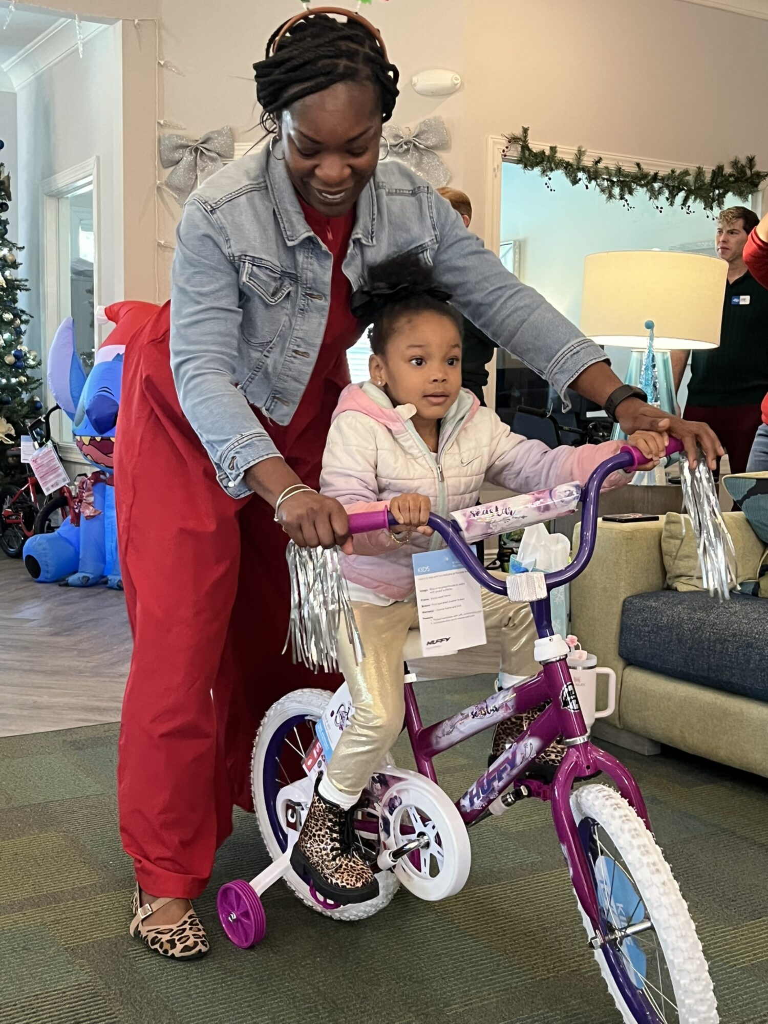 Adult woman helping a young girl ride a purple and white bicycle indoors, with a living room background.