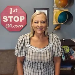 Smiling woman with blonde hair in plaid blouse standing indoors near colorful pipes and a sign.