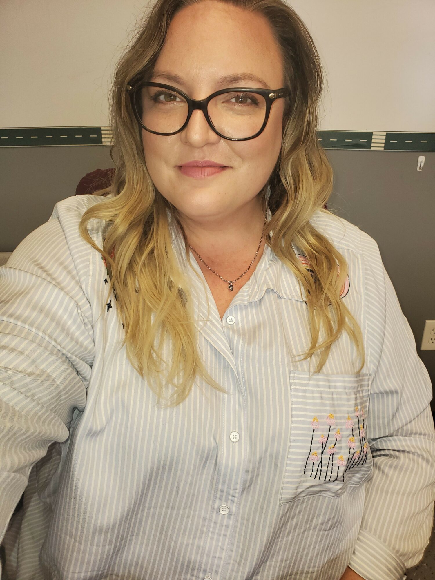 Woman with glasses and wavy hair smiling, wearing a striped shirt, in an indoor setting.
