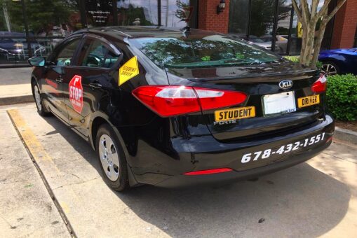 Black Kia sedan parked on sidewalk with student and driver stickers, stop sign, and contact number on rear bumper.