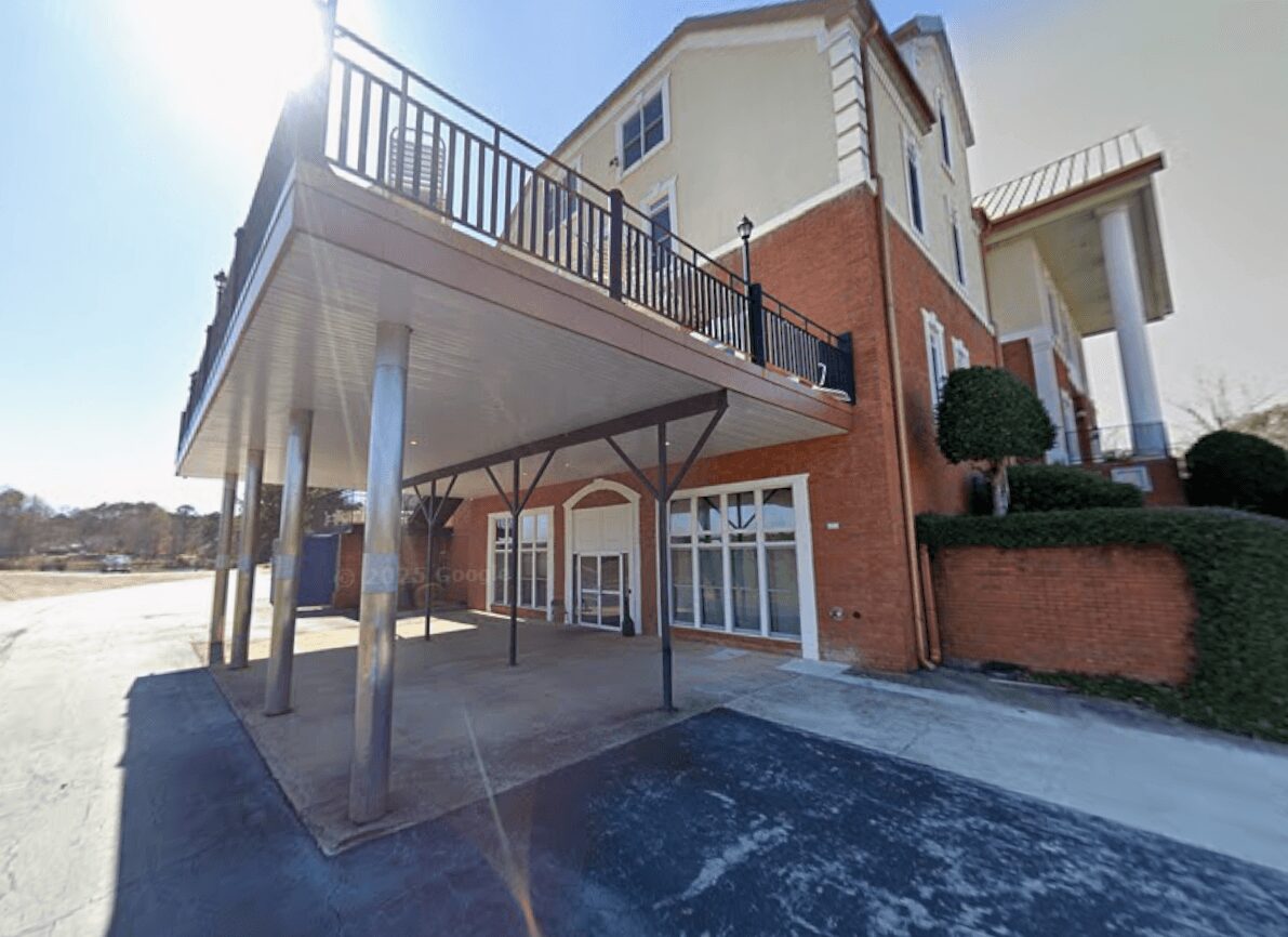 Building with a balcony, support columns, windows, and a brick wall, with a driveway and landscaping nearby.