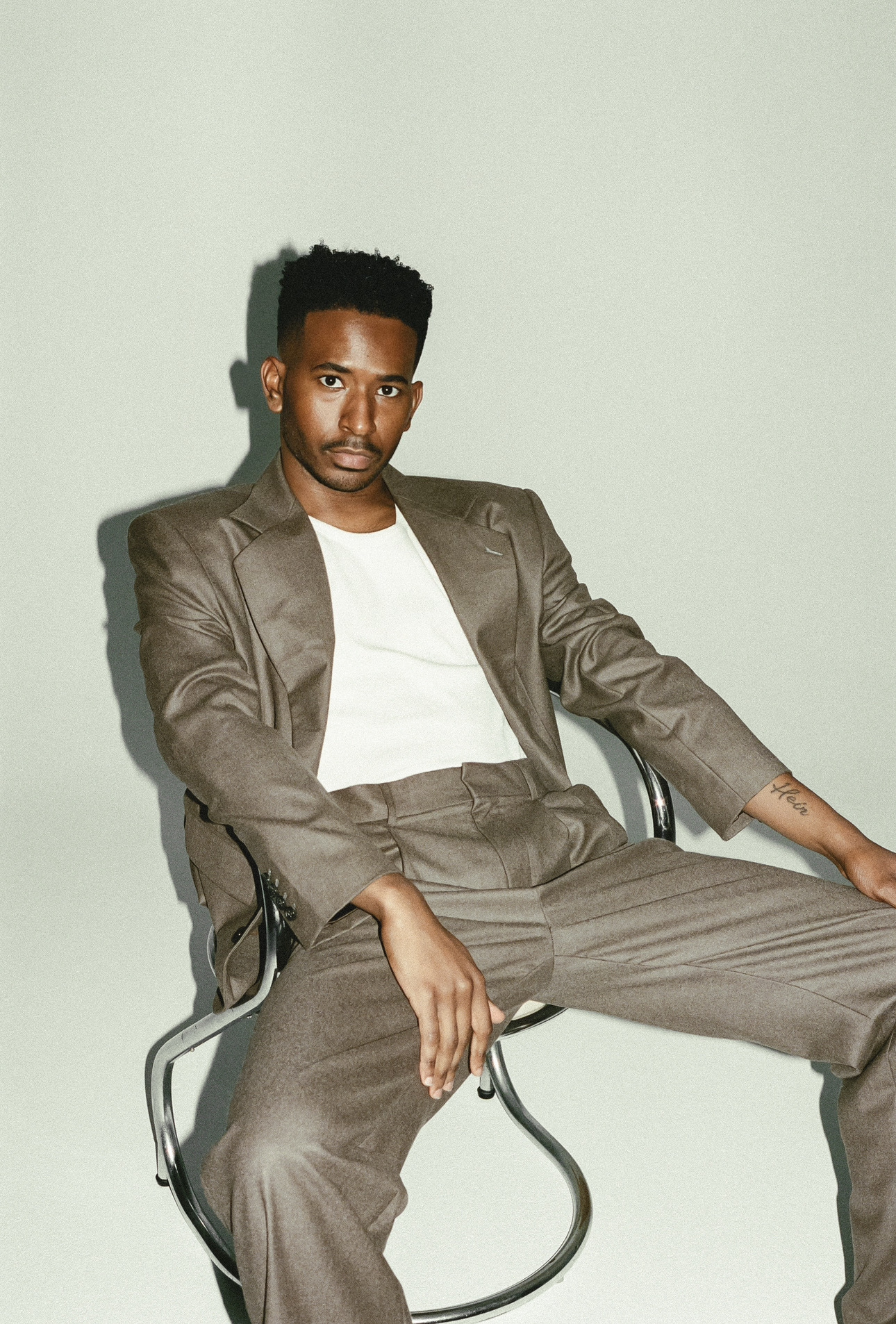 Young man sitting on a chair, wearing a suit and white shirt, against a plain light background.