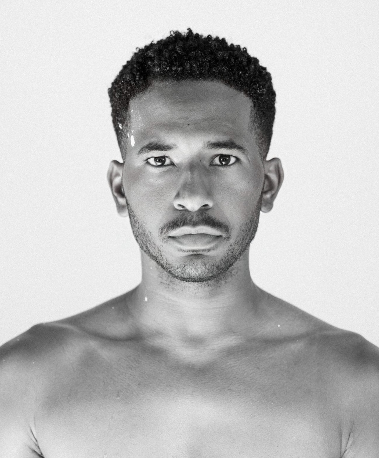 Black and white portrait of a shirtless man with short curly hair, looking directly at the camera.