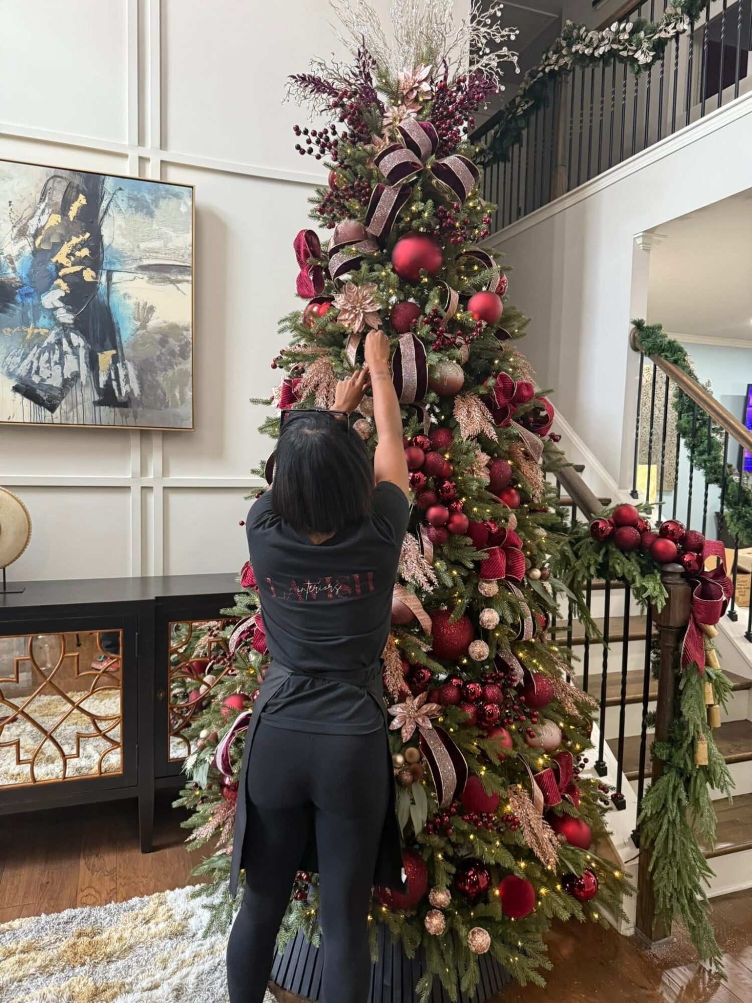 Person decorating a tall Christmas tree with ornaments and ribbons in a decorated indoor space.