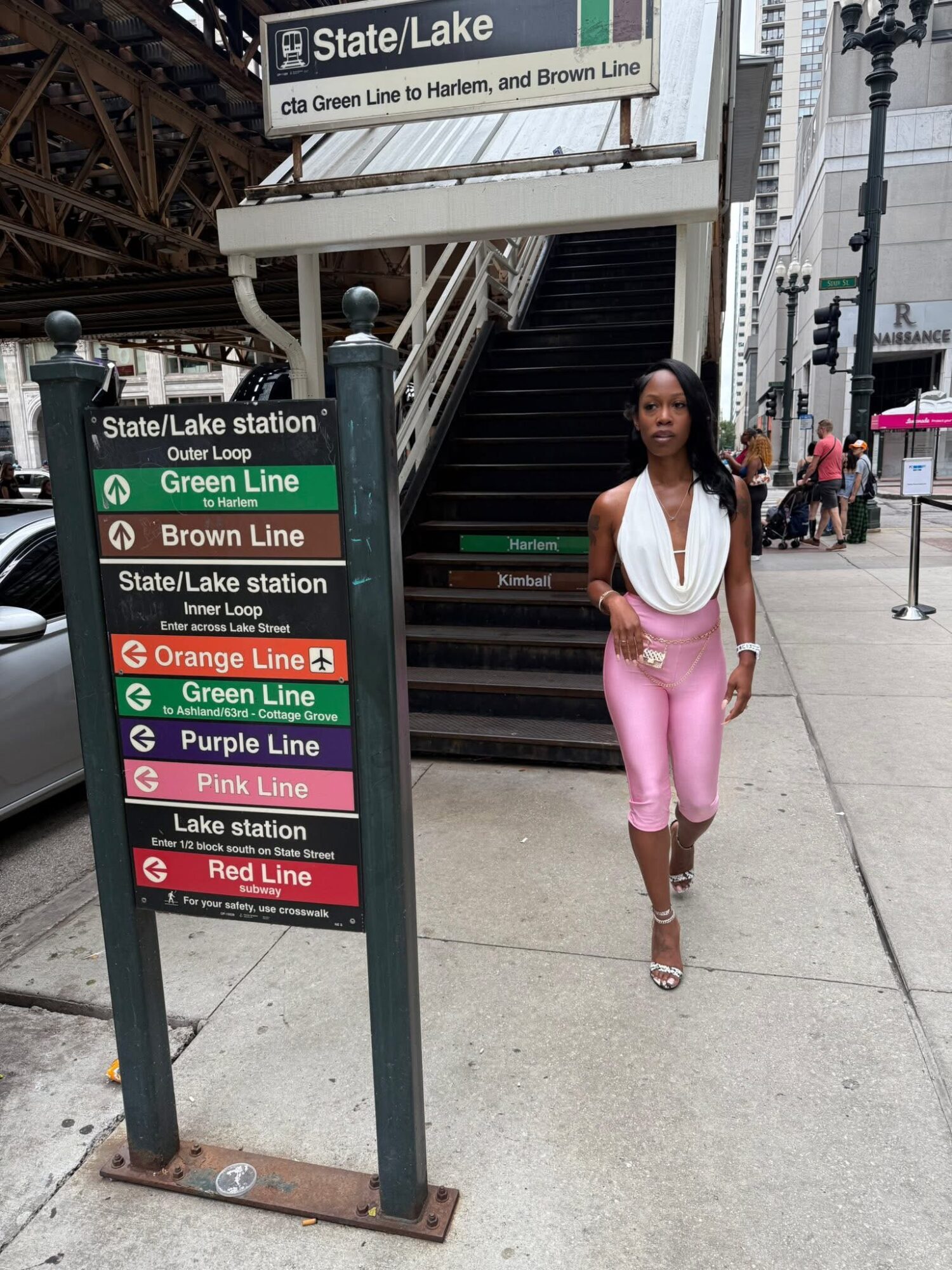 Woman walking on sidewalk near station sign and stairs, city street scene with people and buildings.