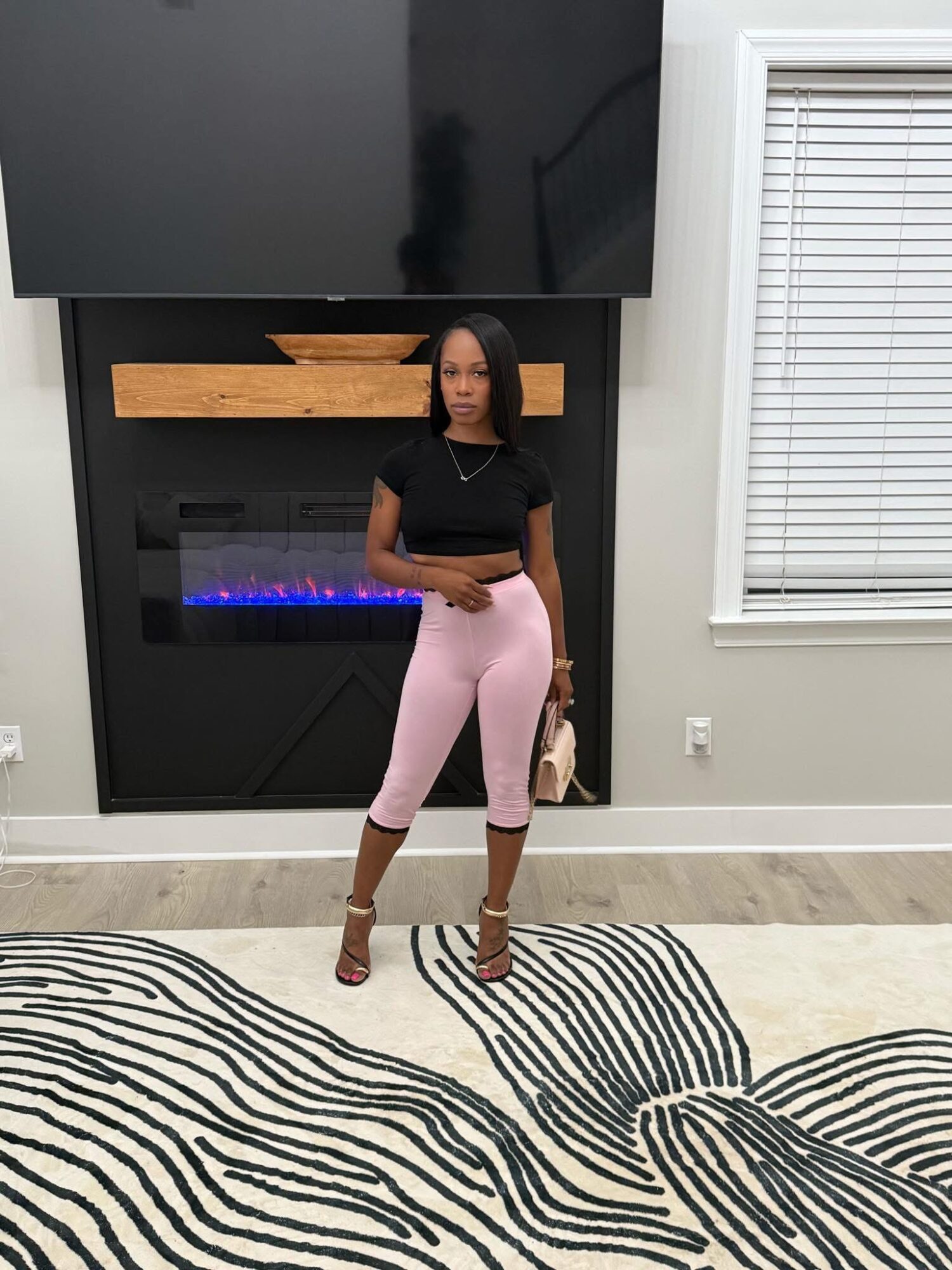 Woman standing in front of a fireplace with a large TV above, wearing pink pants and a black top, holding a bag.