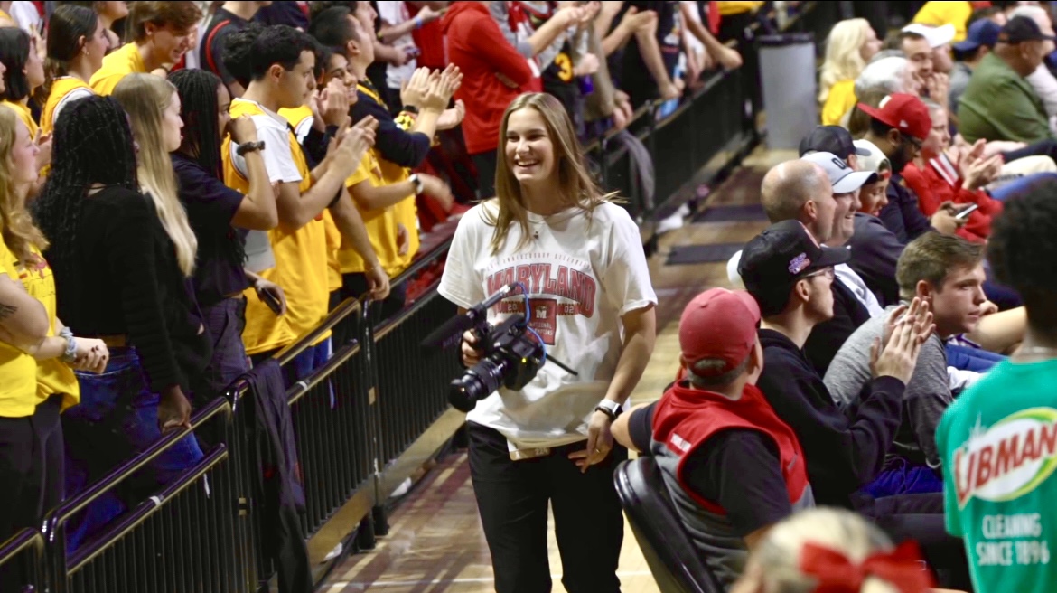 Young woman holding a camera, smiling, surrounded by seated and standing people at an indoor event.