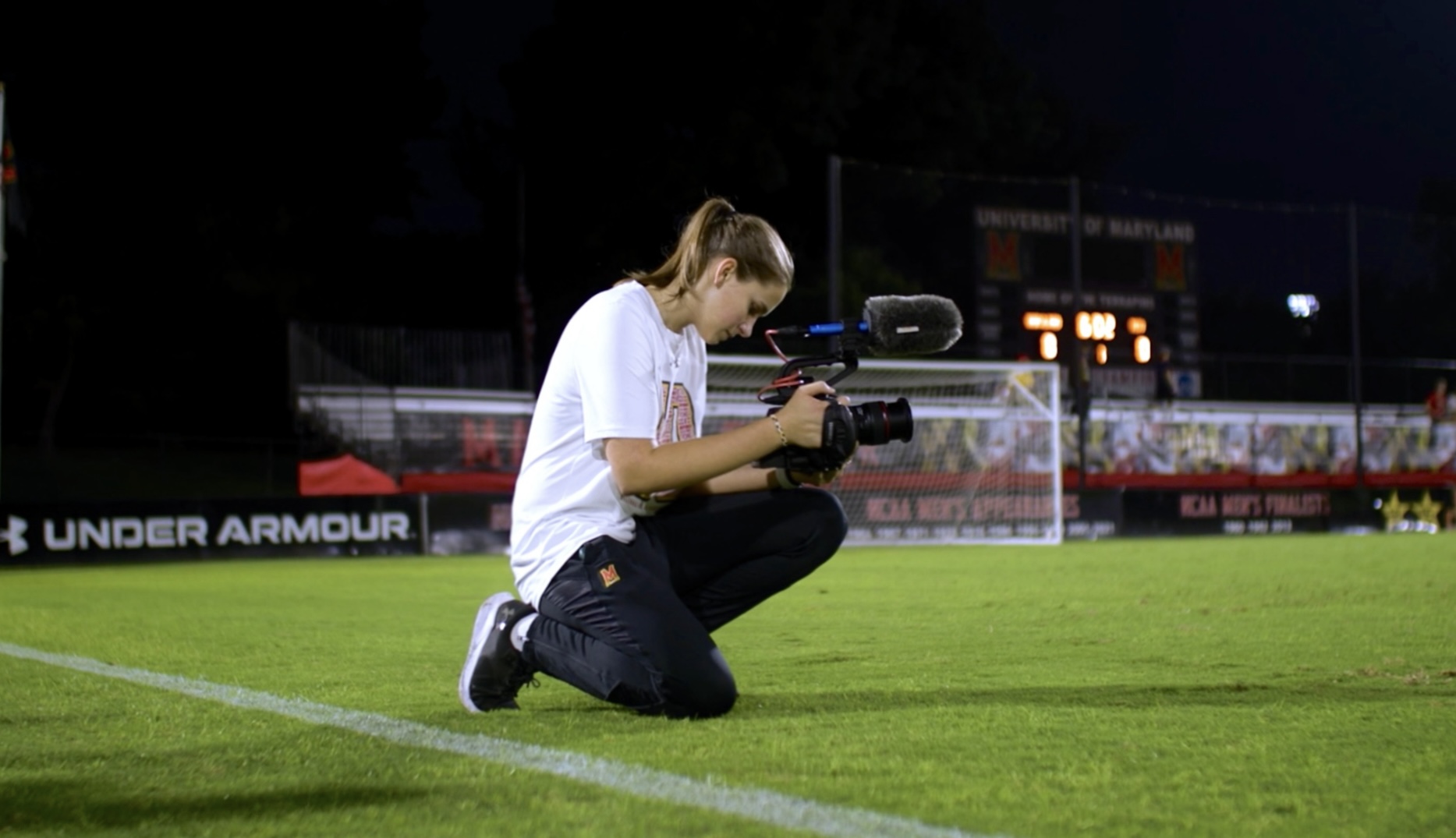 Person kneeling on grass field, filming with professional camera, stadium in background at night.