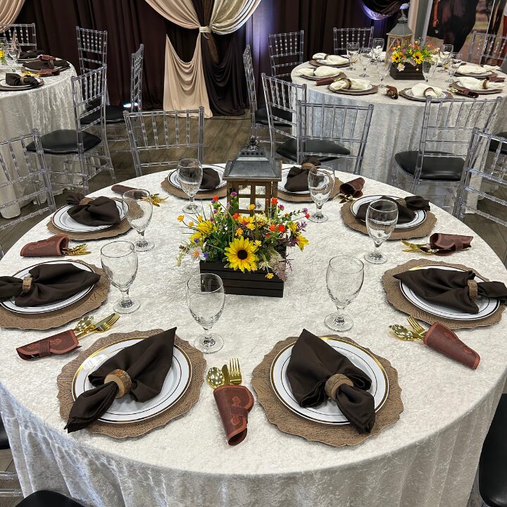 Round table with six place settings, black napkins, gold utensils, floral centerpiece, and glassware, set for a formal event.