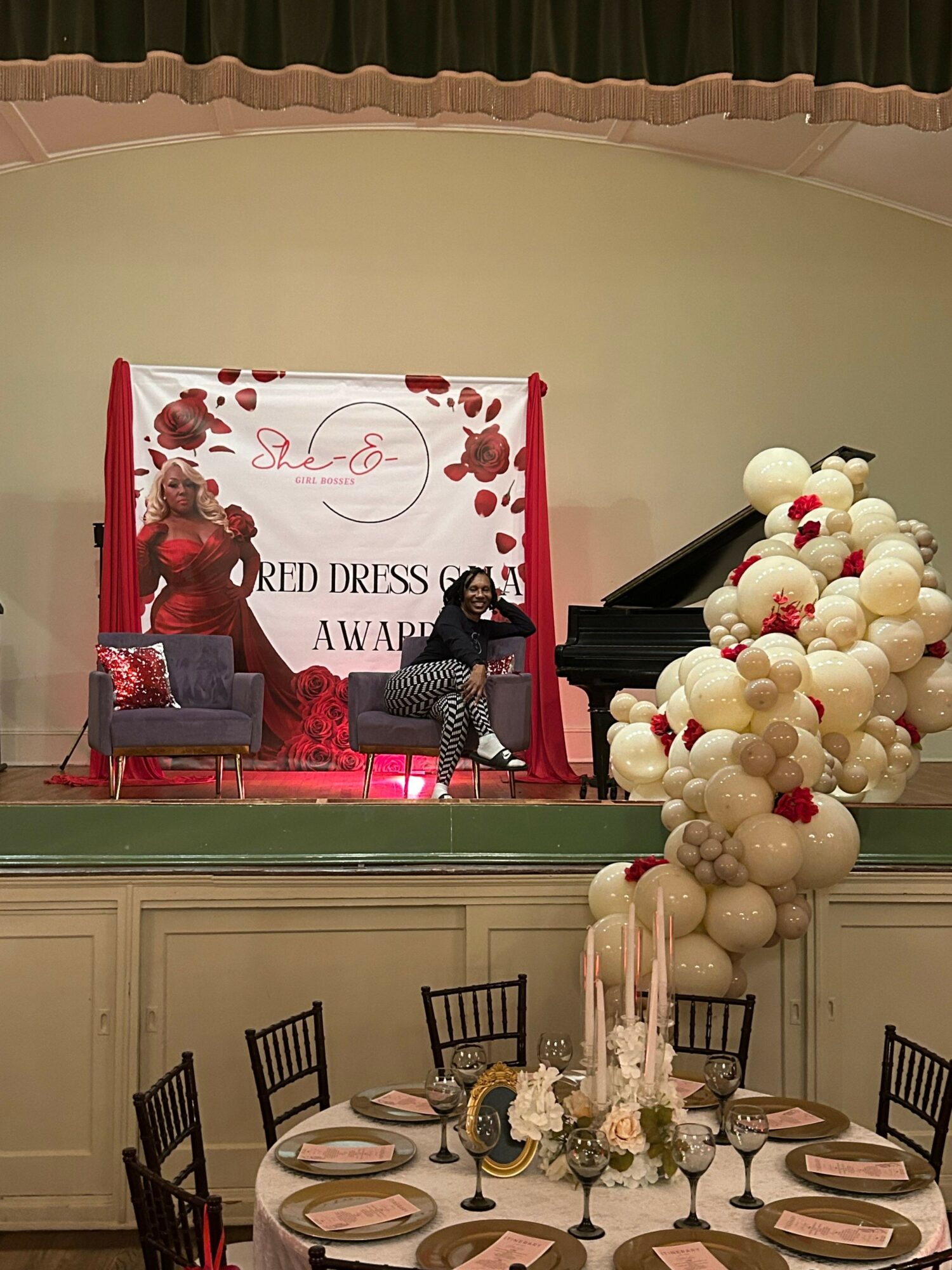 Stage with two women, one standing and one sitting, decorated with a red and white floral backdrop, a purple chair, and a black grand piano. Large white balloon arrangement nearby.