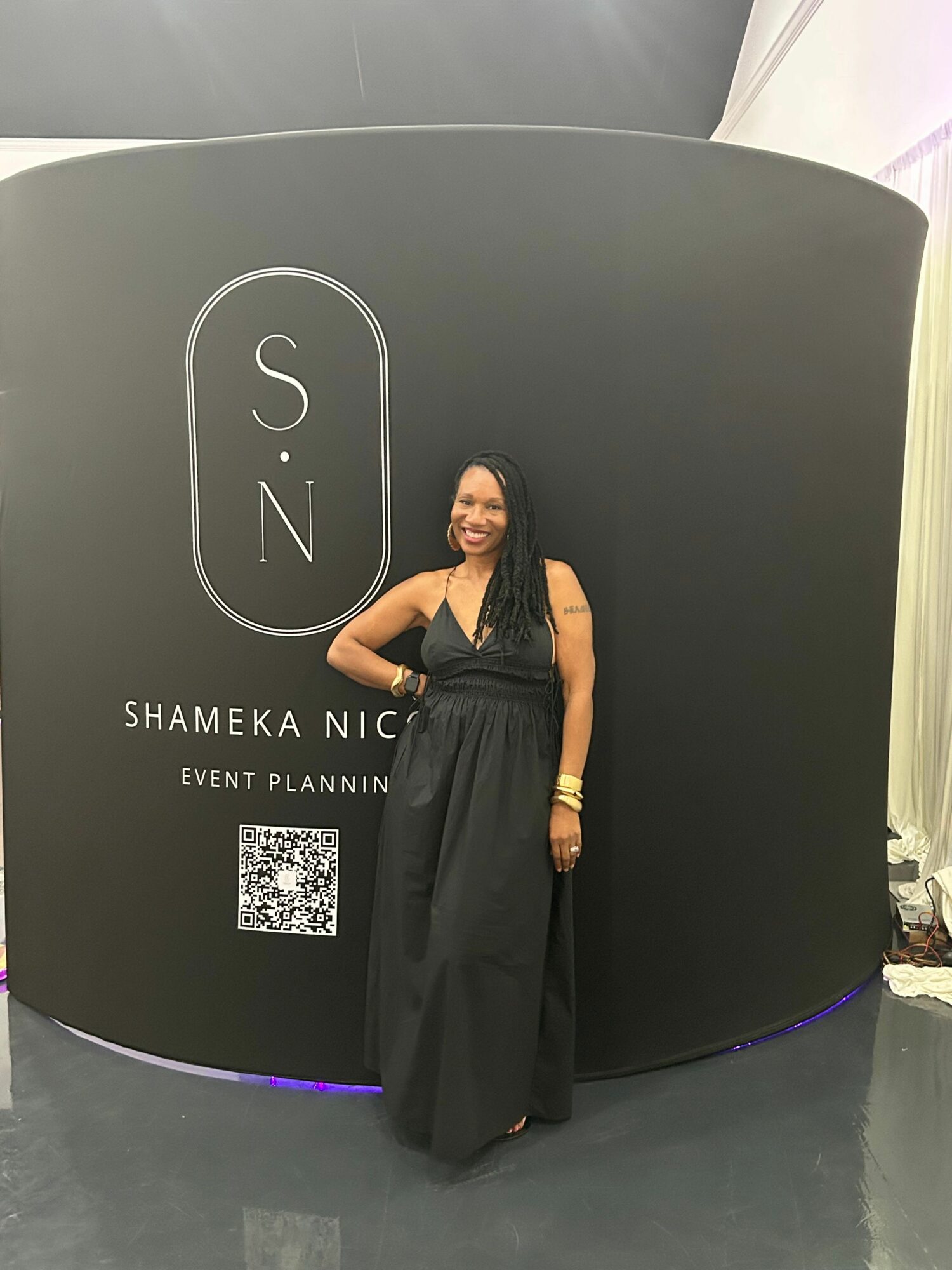 Woman in black dress standing in front of a black backdrop with event branding, smiling, with her hand on her hip.