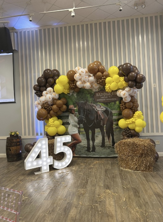 Decorative balloon arch with brown, yellow, and white balloons surrounds a photo backdrop of a person and horse, with a large illuminated '45' sign.