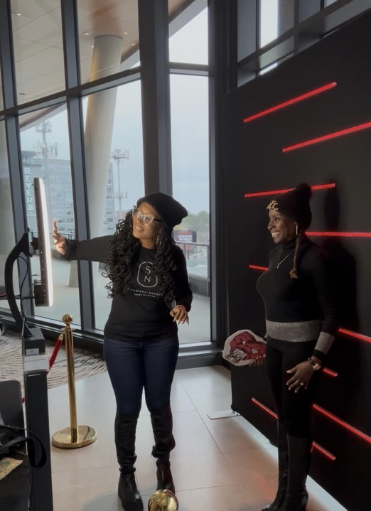 Two women standing indoors near large windows, one taking a selfie, the other smiling, with a black wall with red lines behind them.