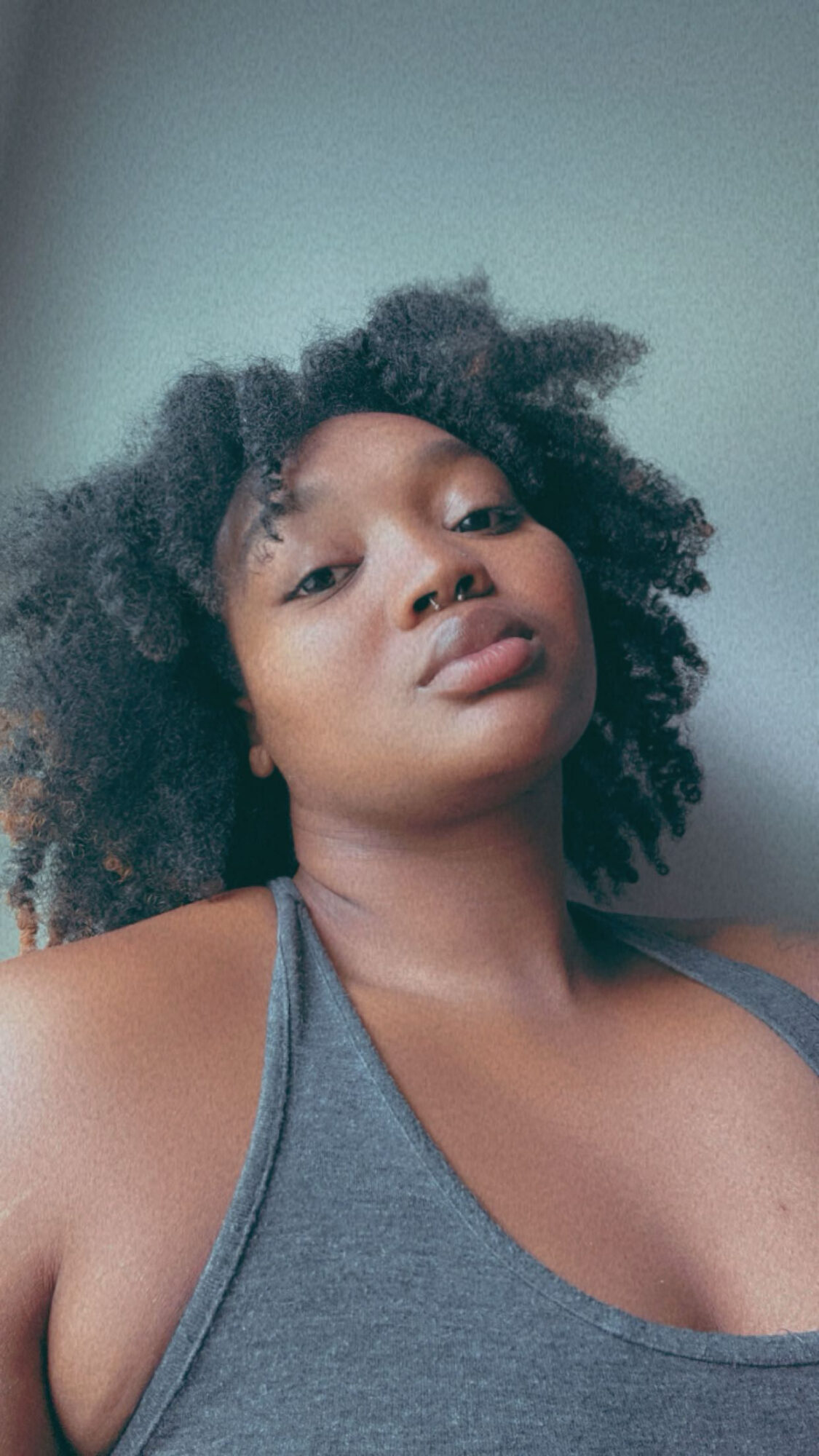 Young woman with natural curly hair wearing a gray tank top, looking at the camera.