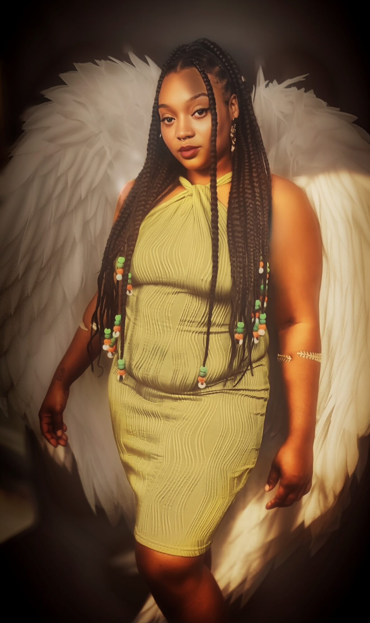 Woman with long braided hair and angel wings, wearing a yellow dress, standing against a dark background.