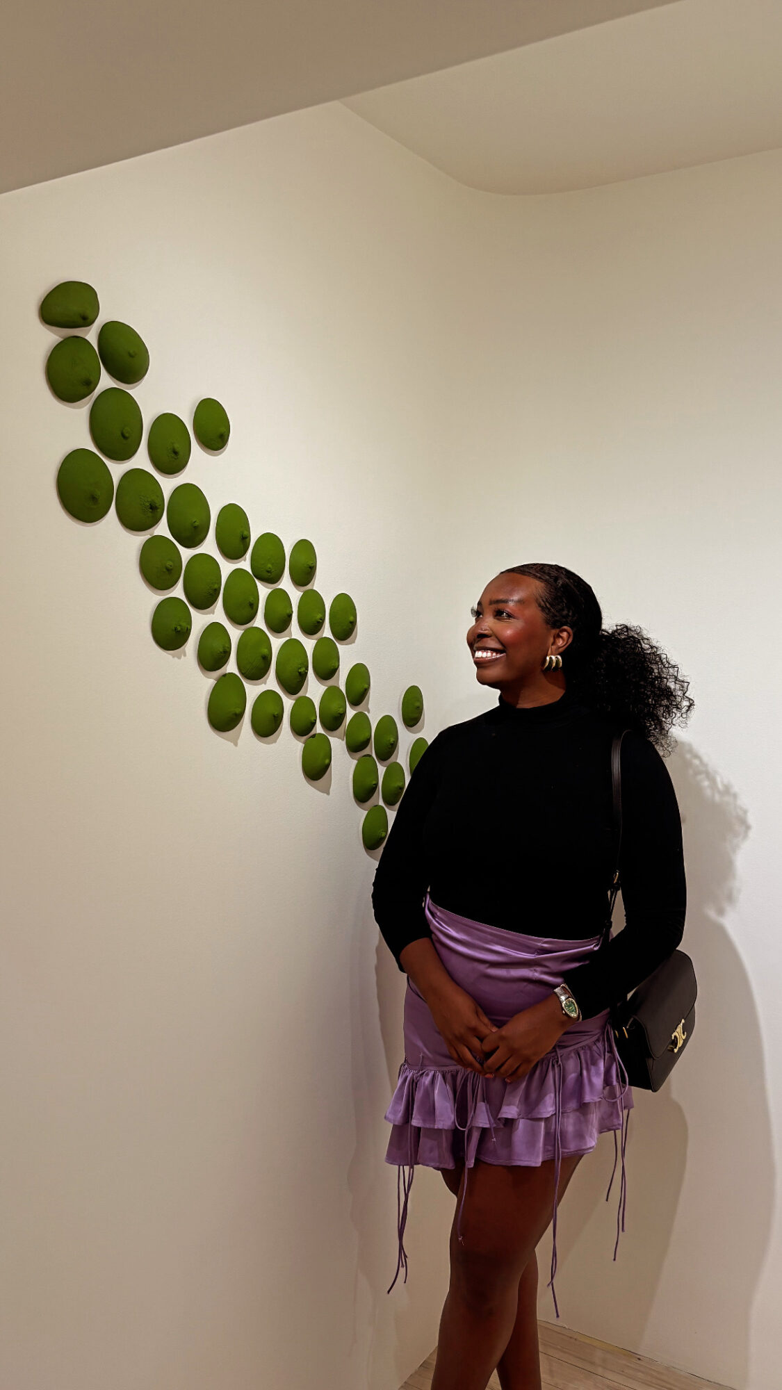 Woman standing indoors near a wall decorated with green circular art pieces, smiling and looking to the side.