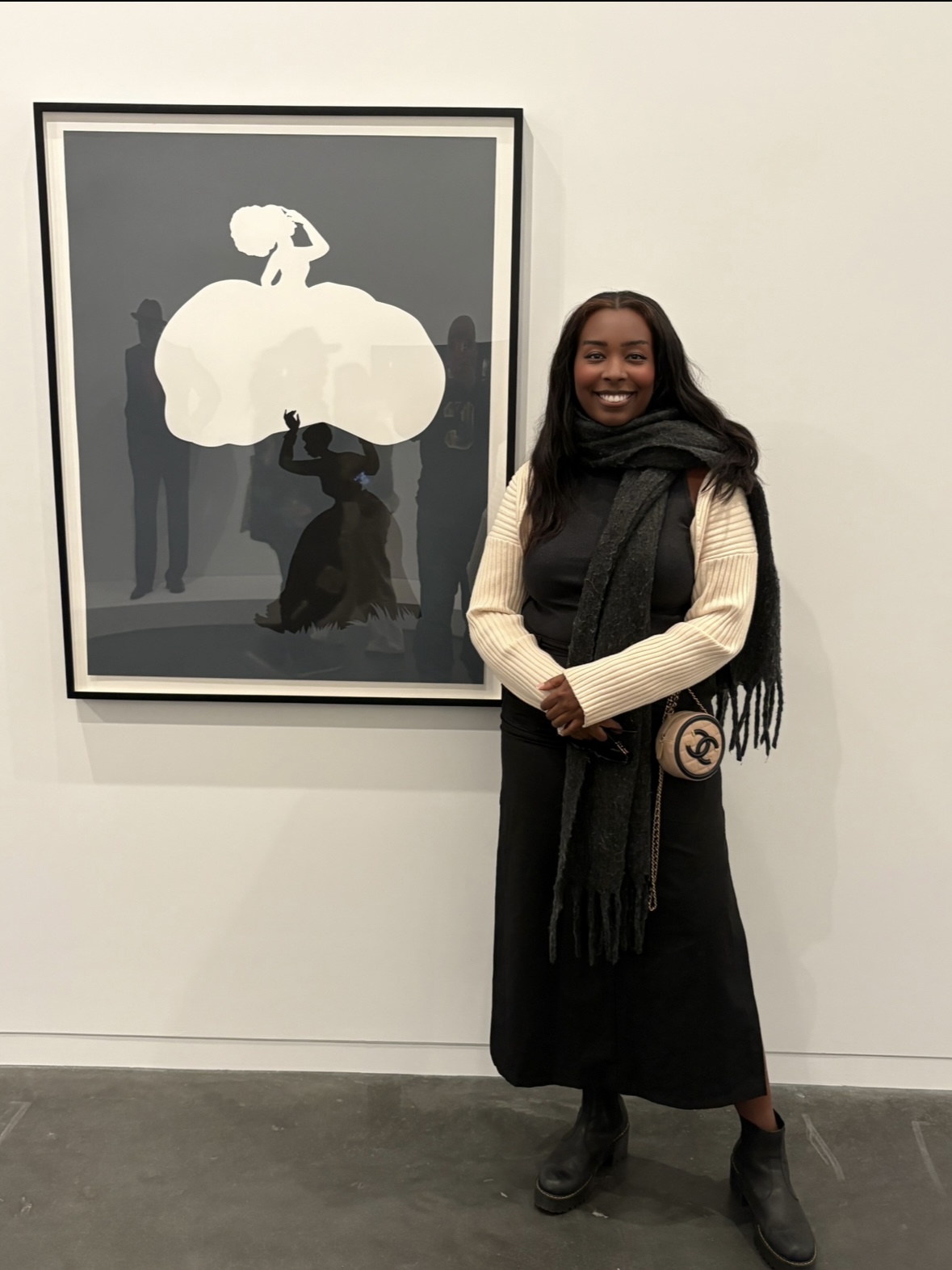 Woman with long dark hair, wearing a beige sweater, black scarf, and black skirt, standing next to framed artwork.