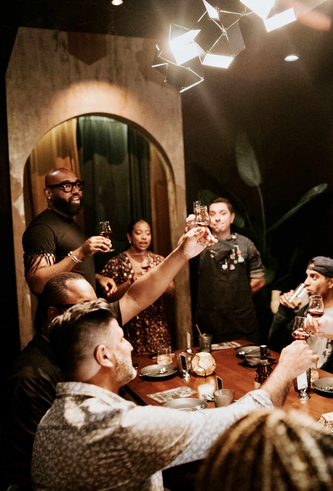 Group of people celebrating around a table, raising glasses in a toast indoors with warm lighting.