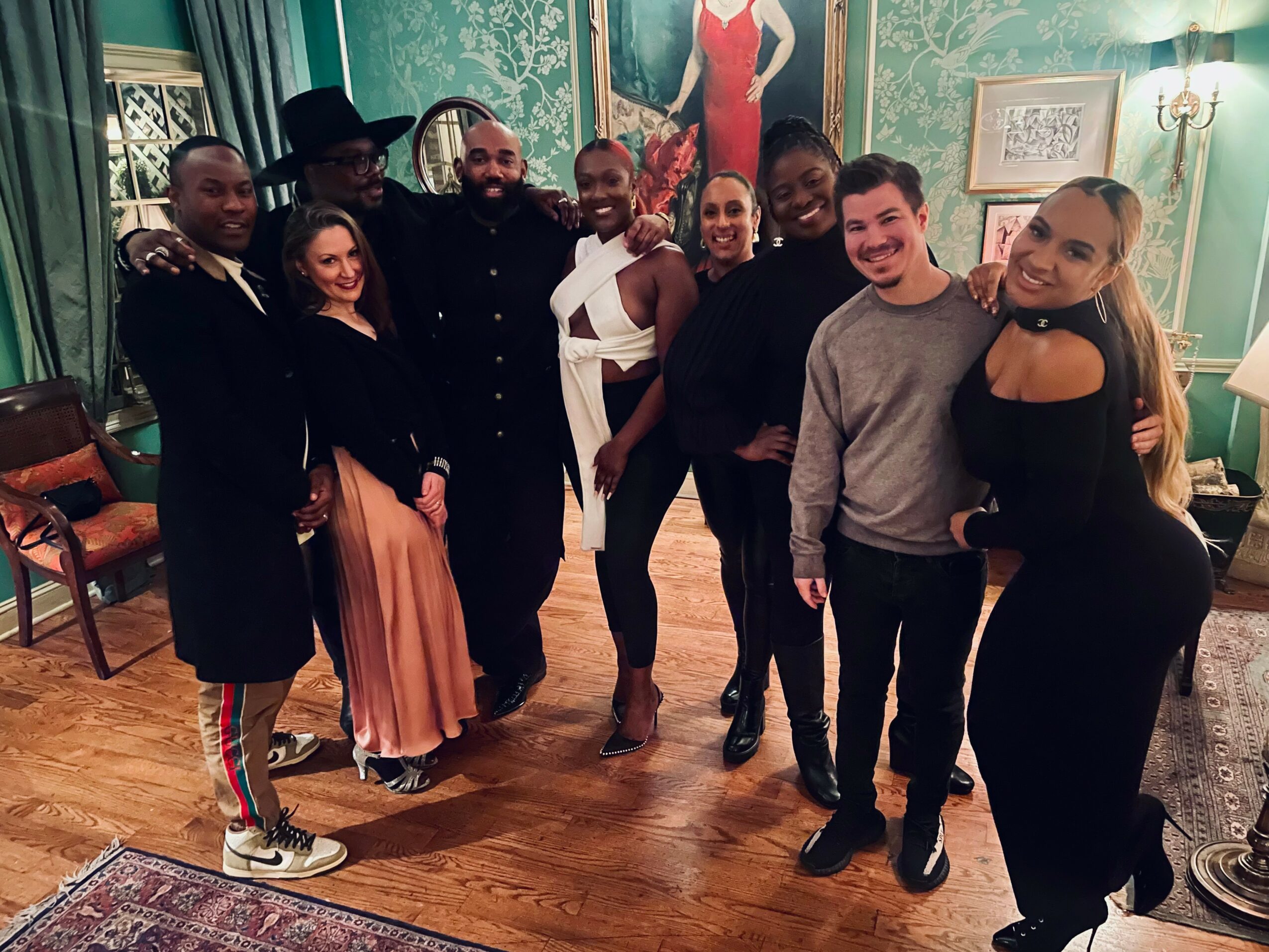 Group of nine people standing together in a room with green walls and wooden floor, smiling for the photo.