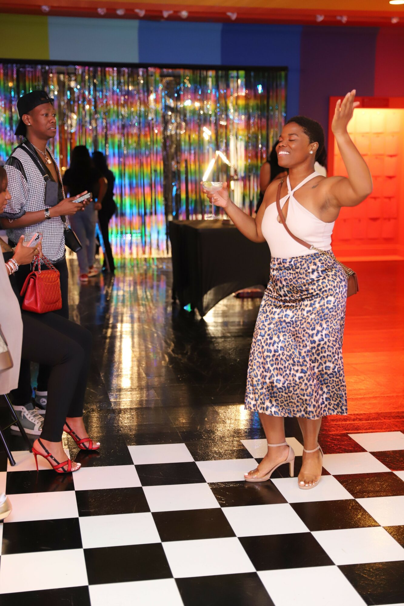 Woman dancing on checkered floor with colorful background and people watching, holding a drink, in a lively indoor setting.