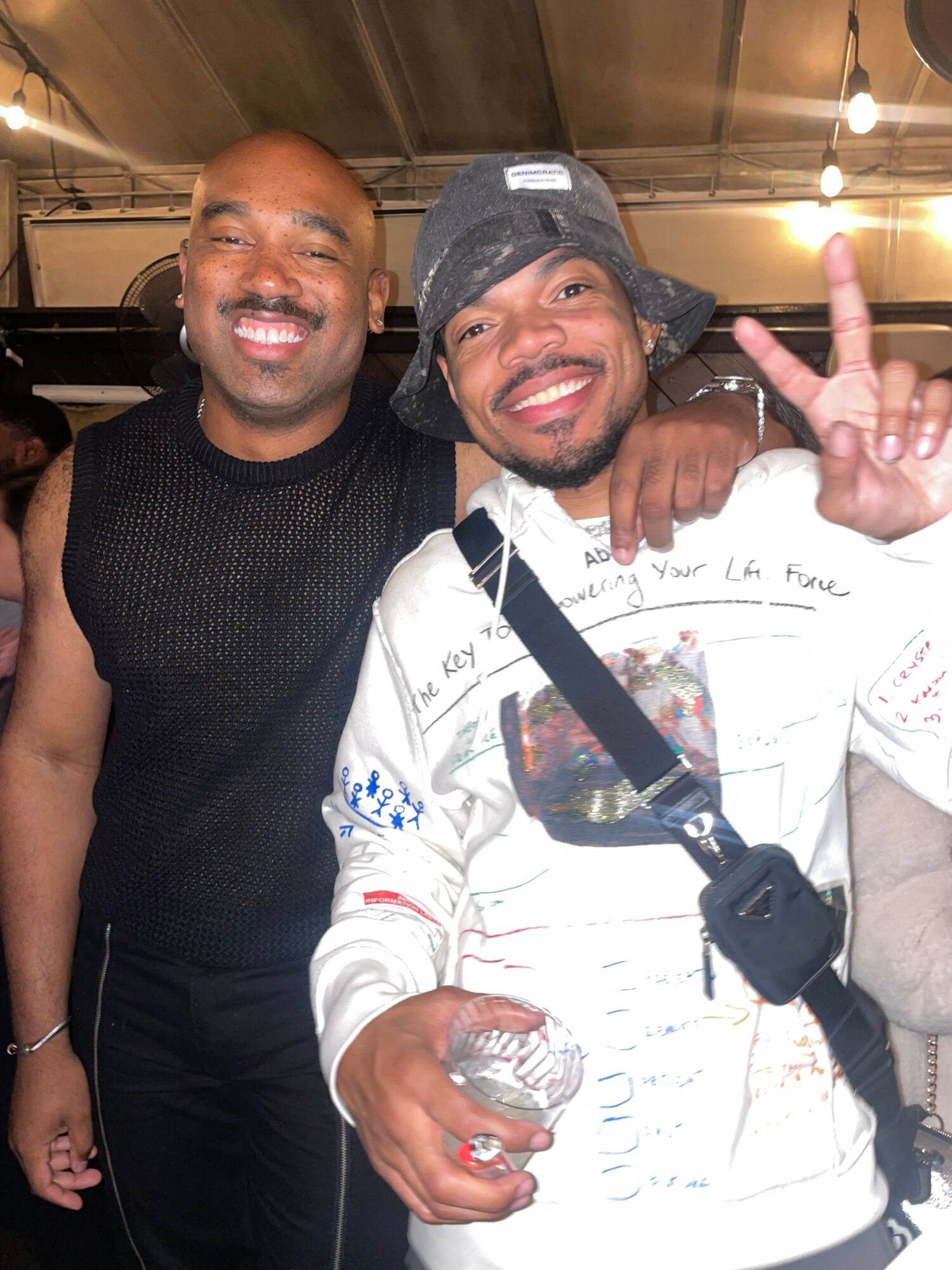 Two men smiling, one making a peace sign, in an indoor setting with warm lighting.