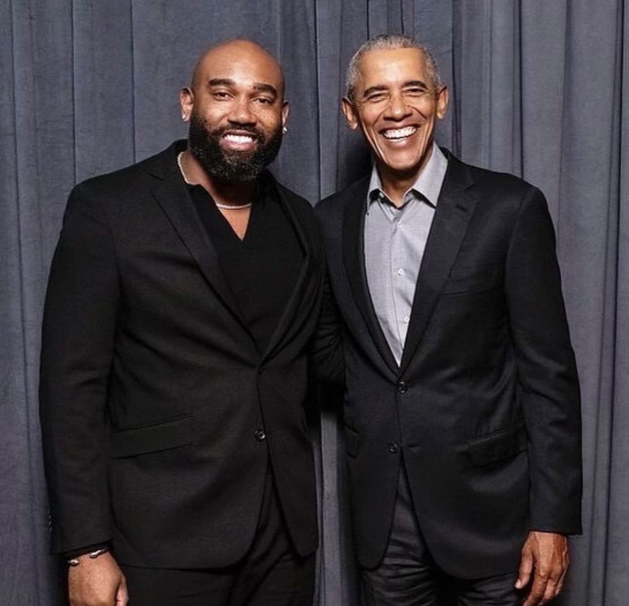 Two men in suits standing side by side, smiling, in front of a gray curtain backdrop.