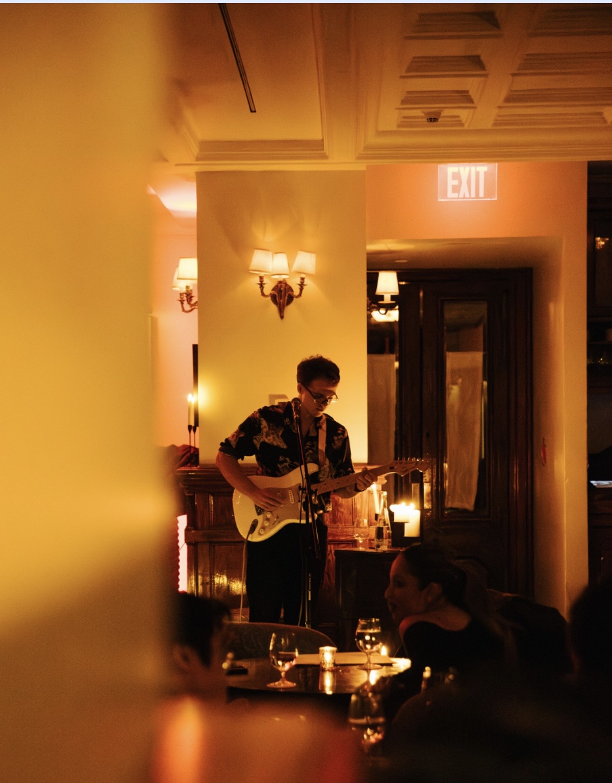 Person playing guitar on stage in a warmly lit room with candles and an exit sign visible.