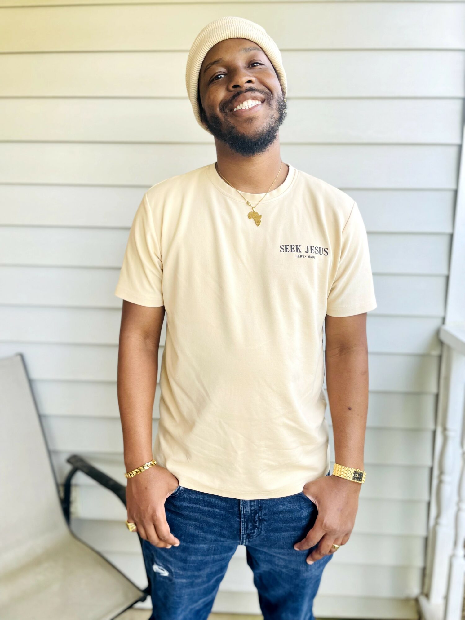 Smiling man wearing a beige beanie, cream T-shirt, and jeans, standing outdoors in front of a light-colored wall.