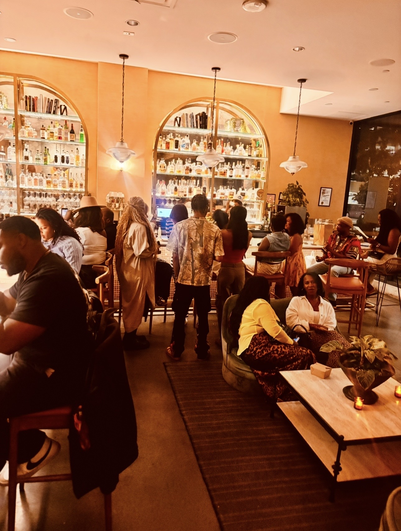 People sit and stand in a warmly lit bar or restaurant with large illuminated shelves of bottles on the wall.