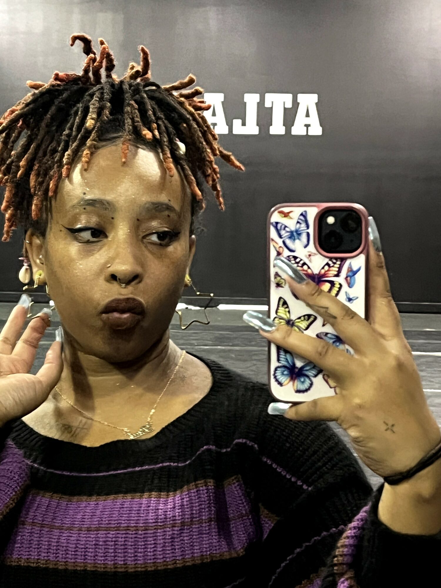 Woman with dreadlocks taking a selfie in mirror, holding phone with colorful case, background with text, wearing dark sweater.