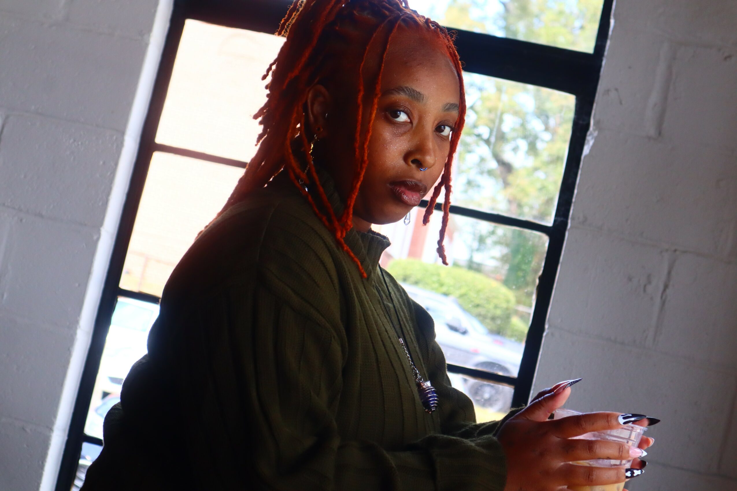 Woman with red dreadlocks looking at camera inside a room with large window and white brick wall.