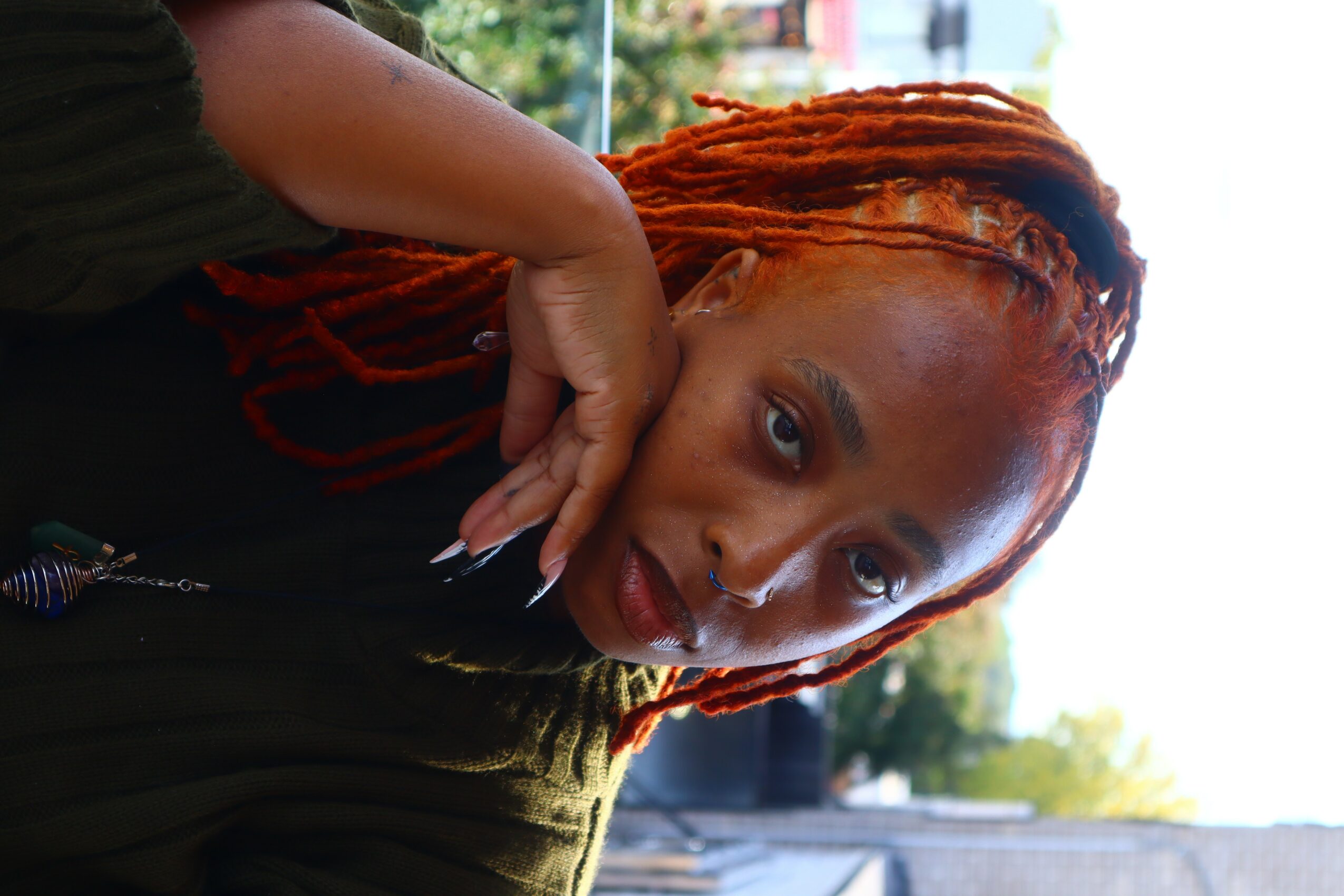 Woman with orange dreadlocks resting her chin on her hand outdoors, looking at the camera.
