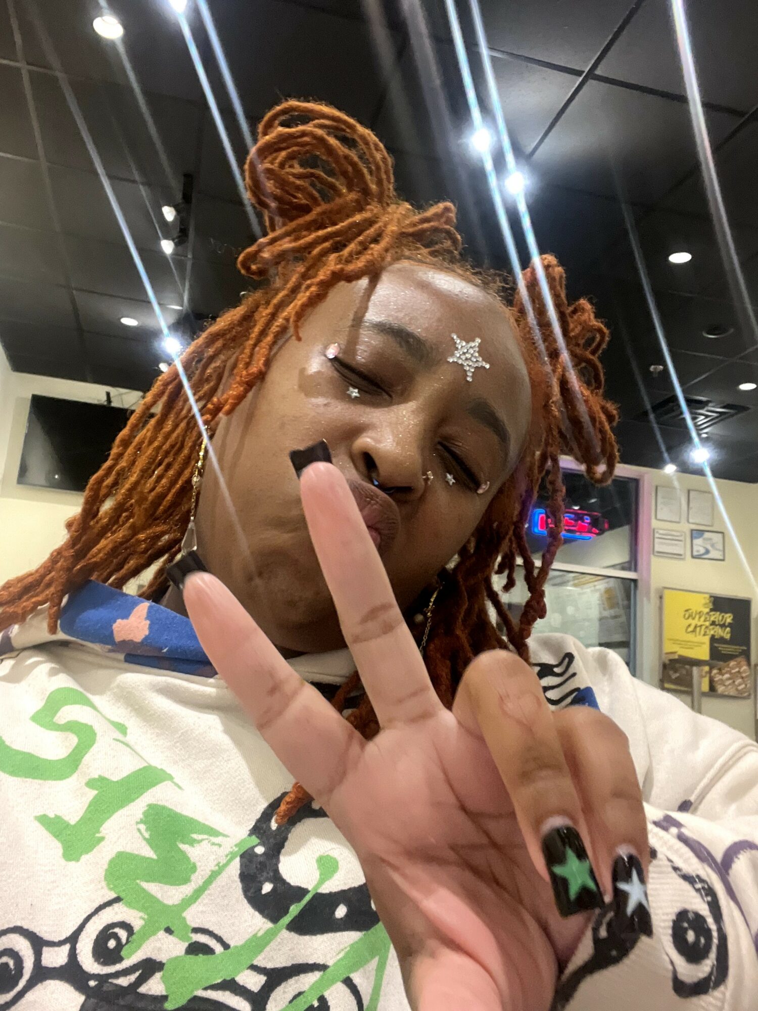 Person with orange dreadlocks making a peace sign, wearing a colorful shirt, in an indoor setting with ceiling lights.