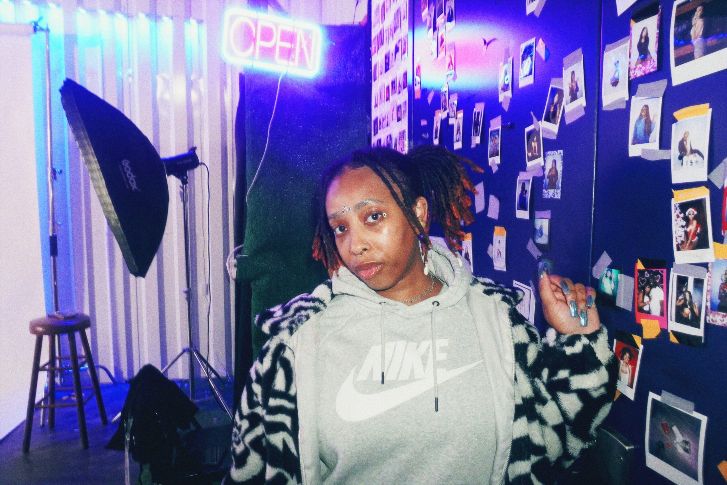 Young woman with dreadlocks in a room decorated with photos and neon lights, wearing a Nike hoodie and patterned jacket.