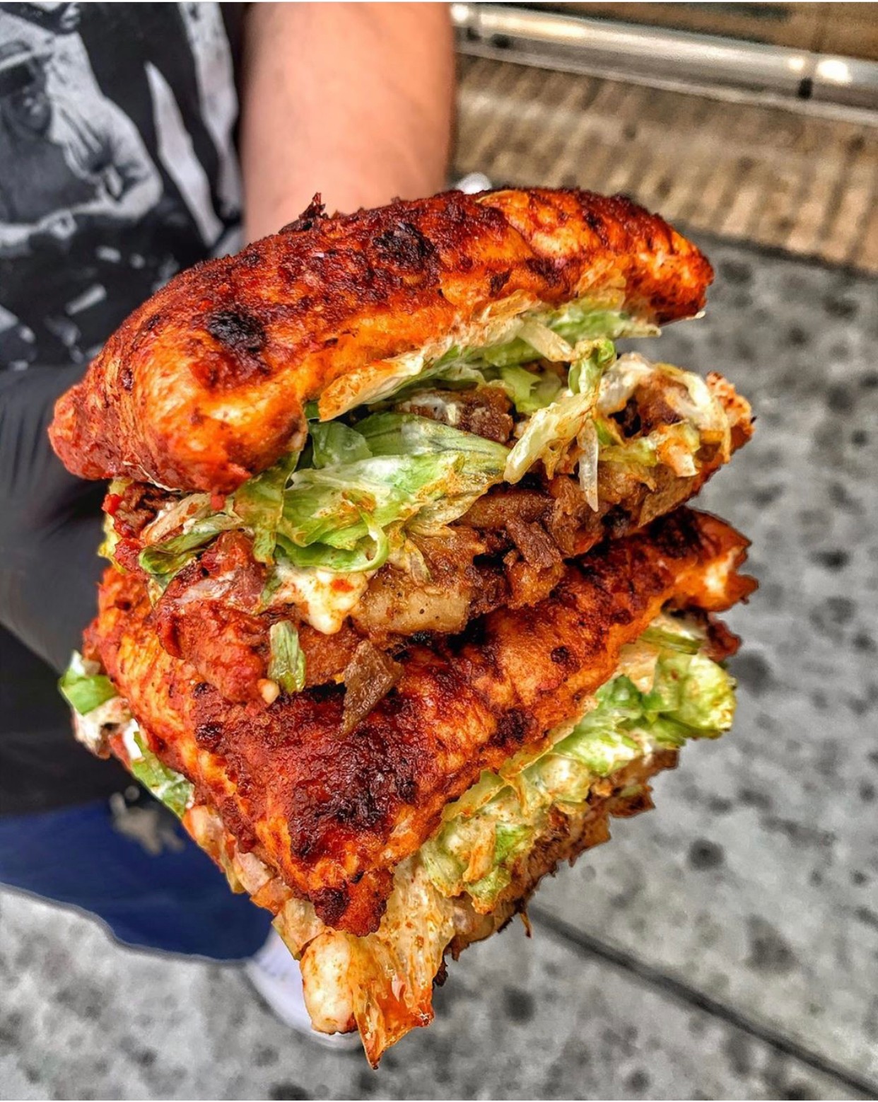 Person holding a large fried chicken sandwich with lettuce, pickles, and sauce, outdoors on a concrete surface.