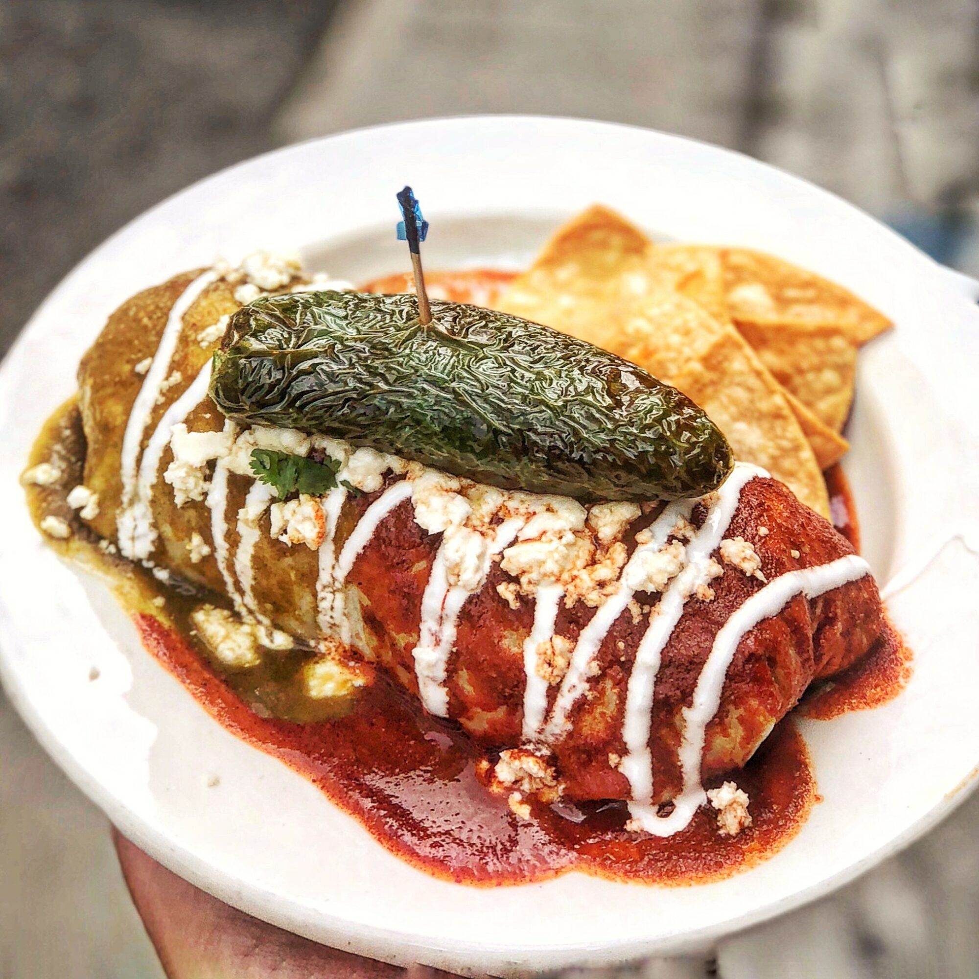 Plate with a stuffed chili pepper covered in sauce, topped with cheese and a pickle, with chips on the side.