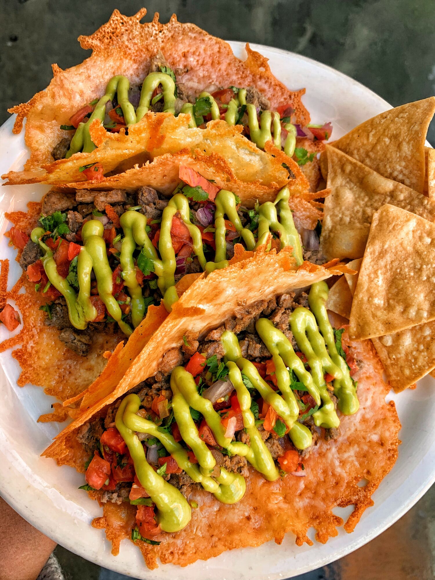 Three tacos with shredded meat, green sauce, and crispy shells on a white plate, with fried chips on the side.