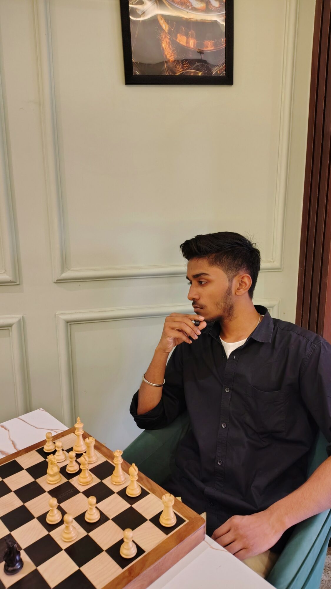 Young man with dark hair and beard, wearing a black shirt, sitting at a chessboard in a room with cream-colored walls.