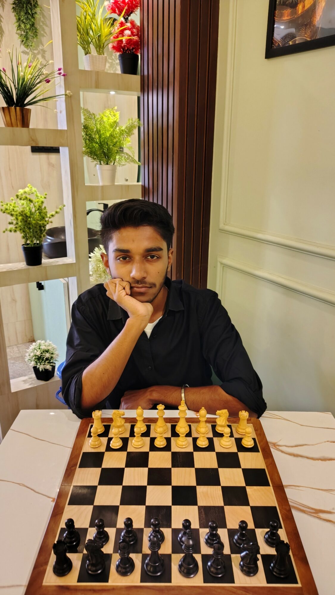 Young man sitting at a table with a chessboard, resting his chin on his hand, in a room with plants and wooden decor.