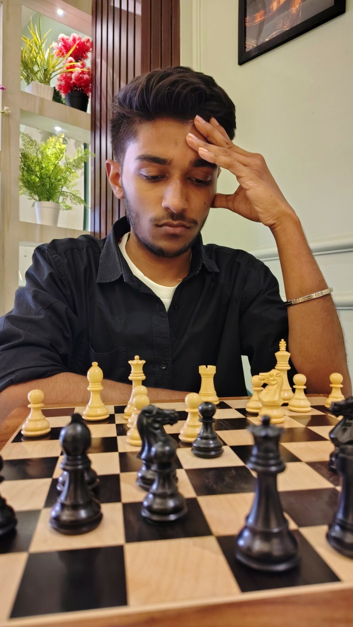 Young man concentrating on chess game, hand on forehead, in indoor setting with plants and framed picture.