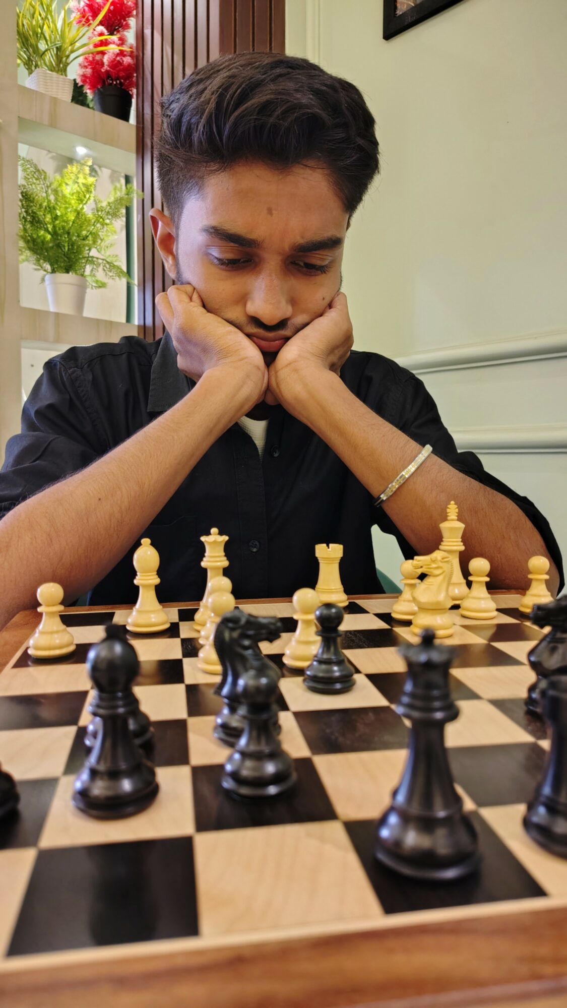 Young person concentrating on a chess game, resting their chin on hands, in a room with plants and framed pictures.