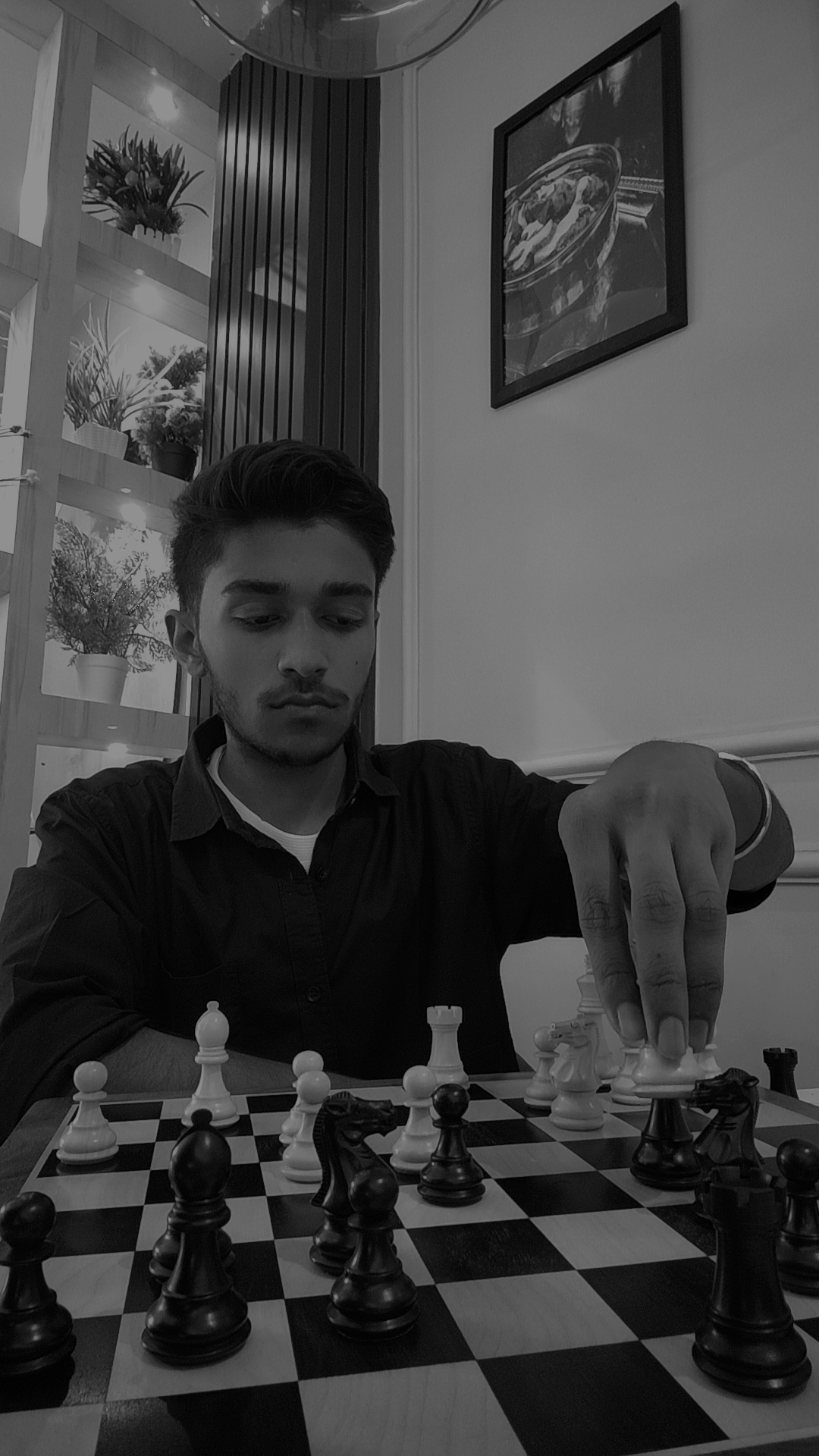 Young man playing chess at a table indoors, with plants and framed picture on the wall behind him.