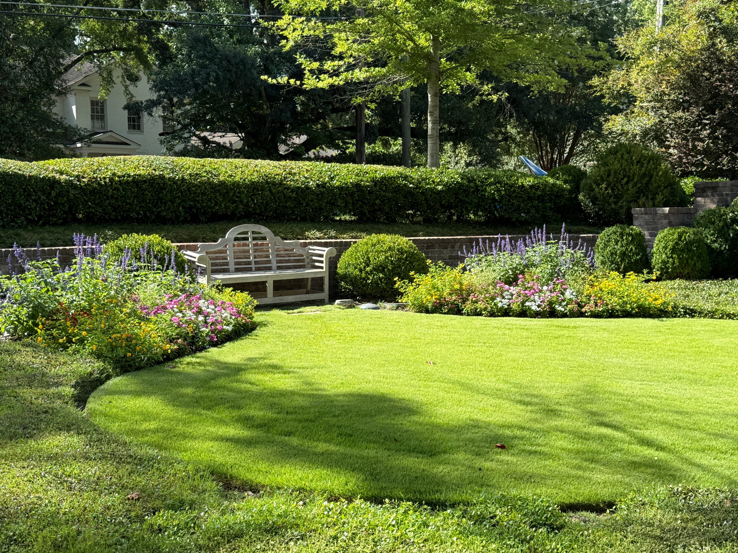 Well-maintained garden with a white bench, colorful flower beds, green grass, and trees in the background.