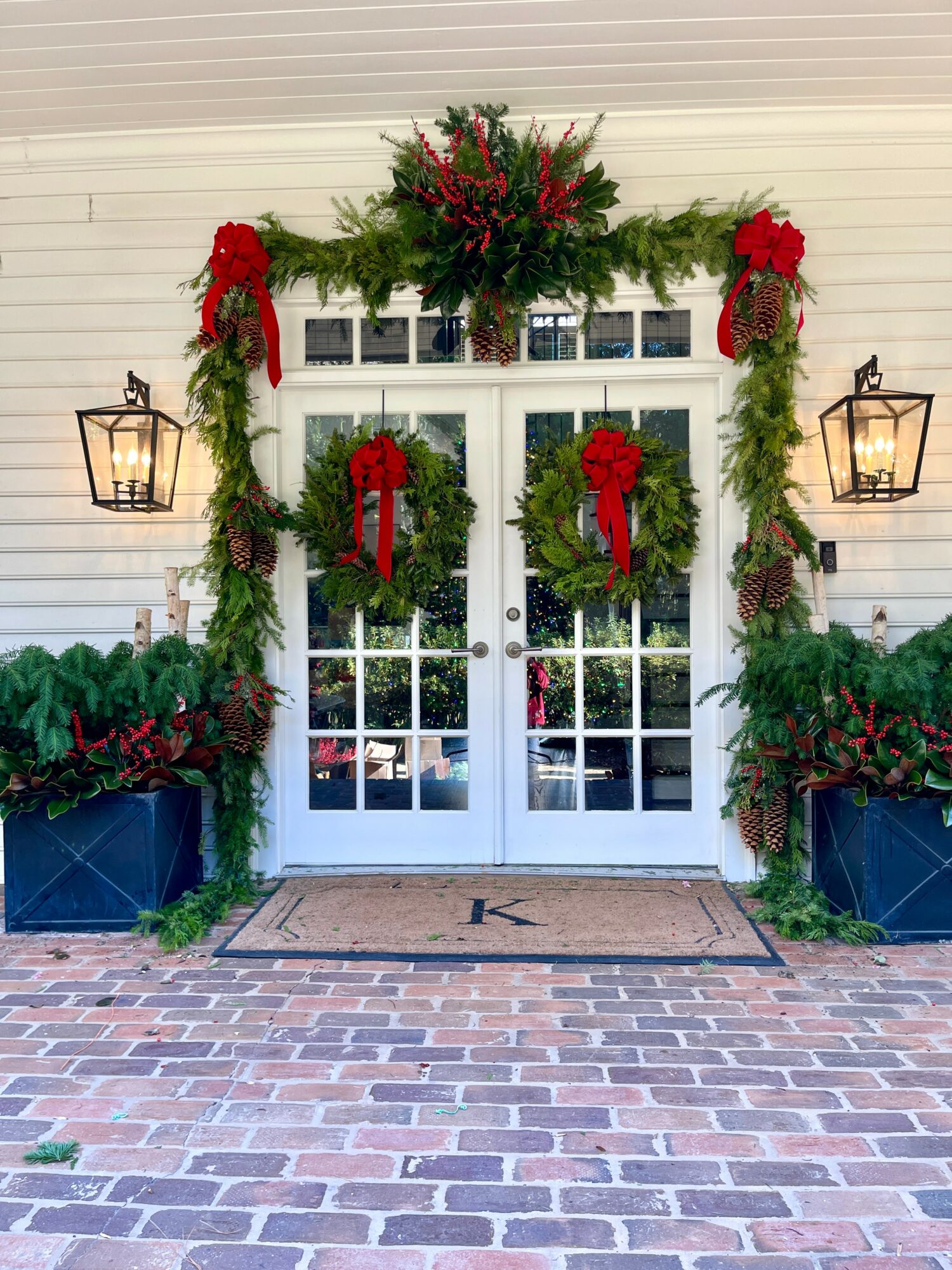 White double doors decorated with red bows and greenery, flanked by lanterns and potted plants, with brick pavement in front.