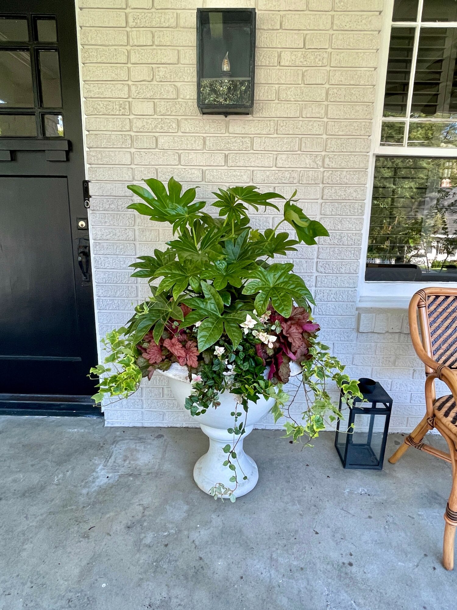 Large floral arrangement in a white vase with green and purple leaves, white flowers, placed outside near a brick wall.
