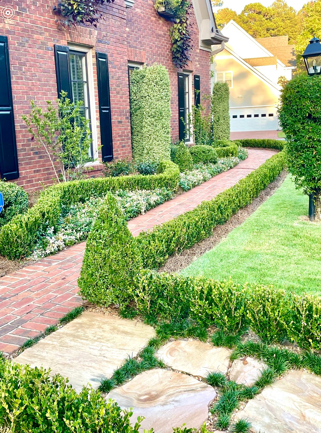 Brick house with tall bushes, a brick pathway, and a well-maintained garden with trimmed hedges and trees.