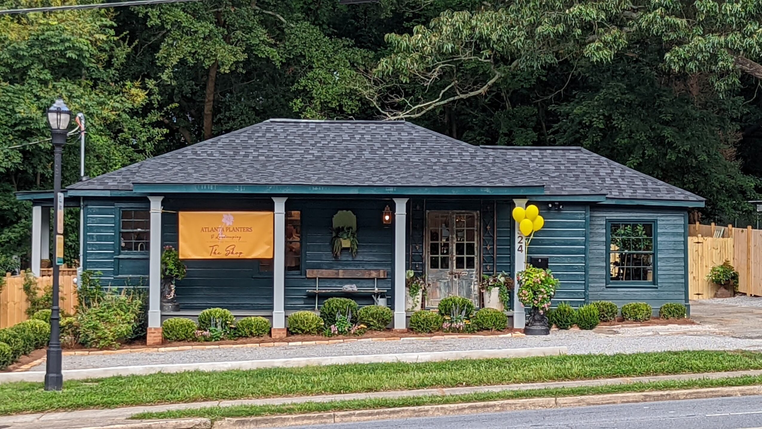 Single-story blue building with porch, yellow balloons, and surrounding greenery, situated along a street with a sidewalk.